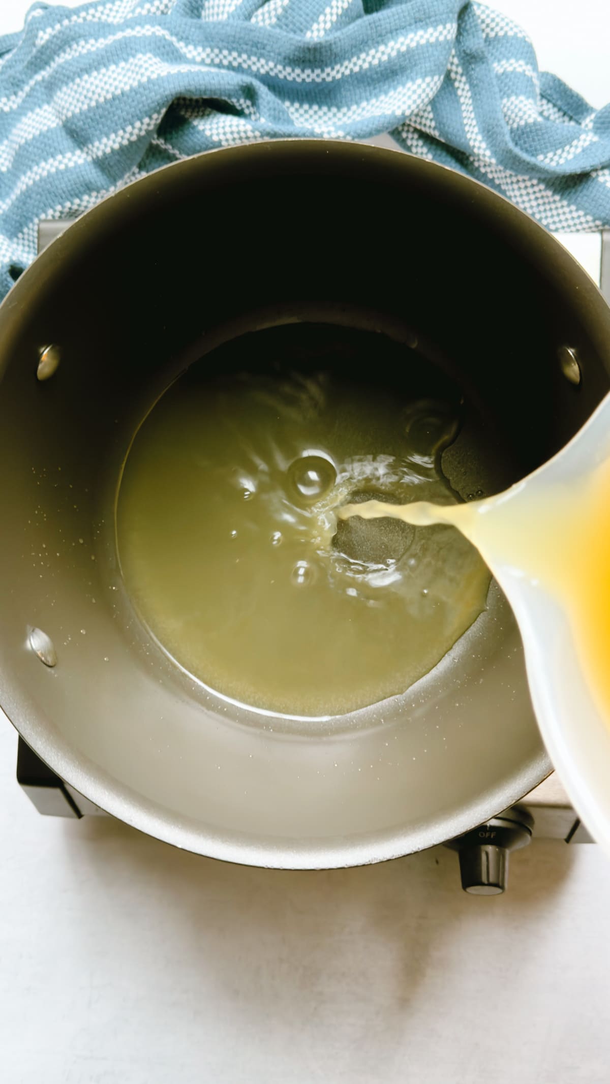 Pouring chicken broth into a pot on top of a gray surface, with a blue napkin on the side.