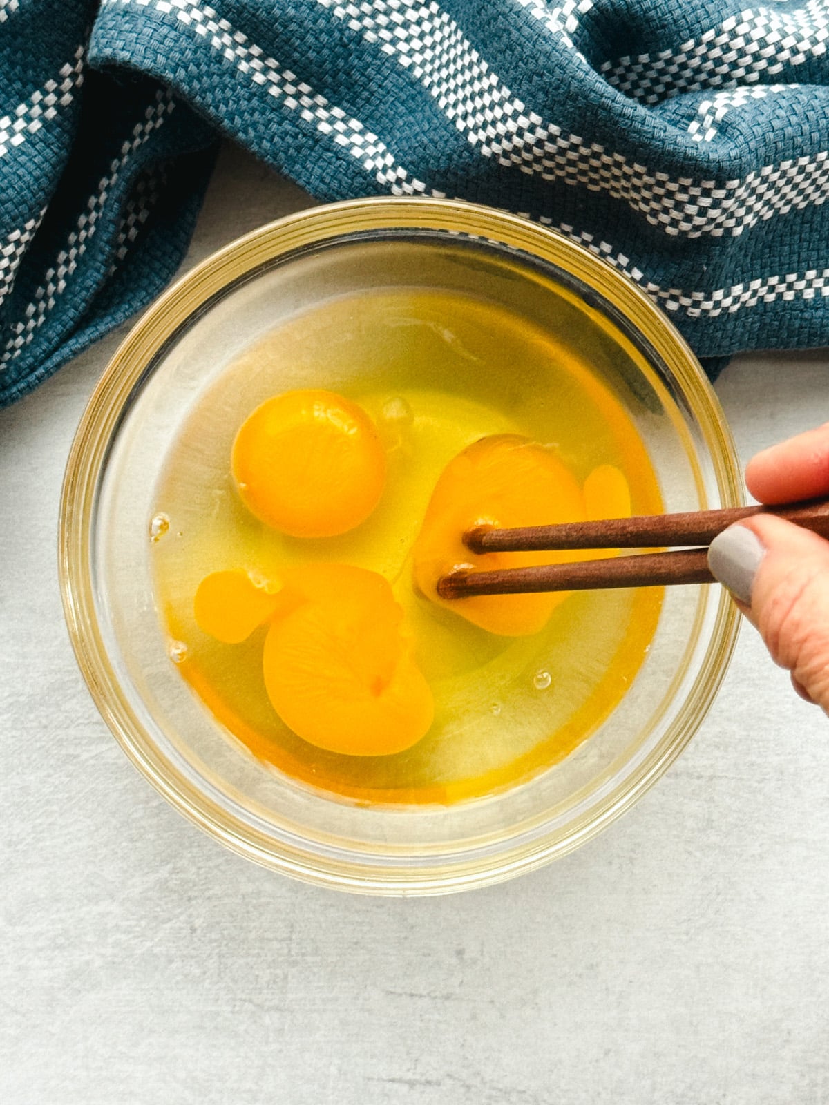 Three eggs in a glass bowl being stirred with chopsticks on top of a gray surface with a blue napkin on the side.