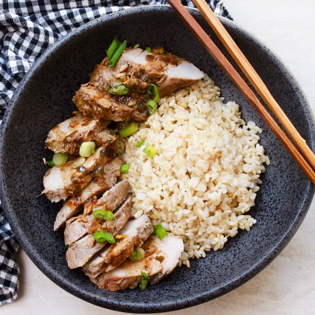 Chinese five spice pork tenderloin slices in a black bowl with brown rice and a pair of chopsticks.