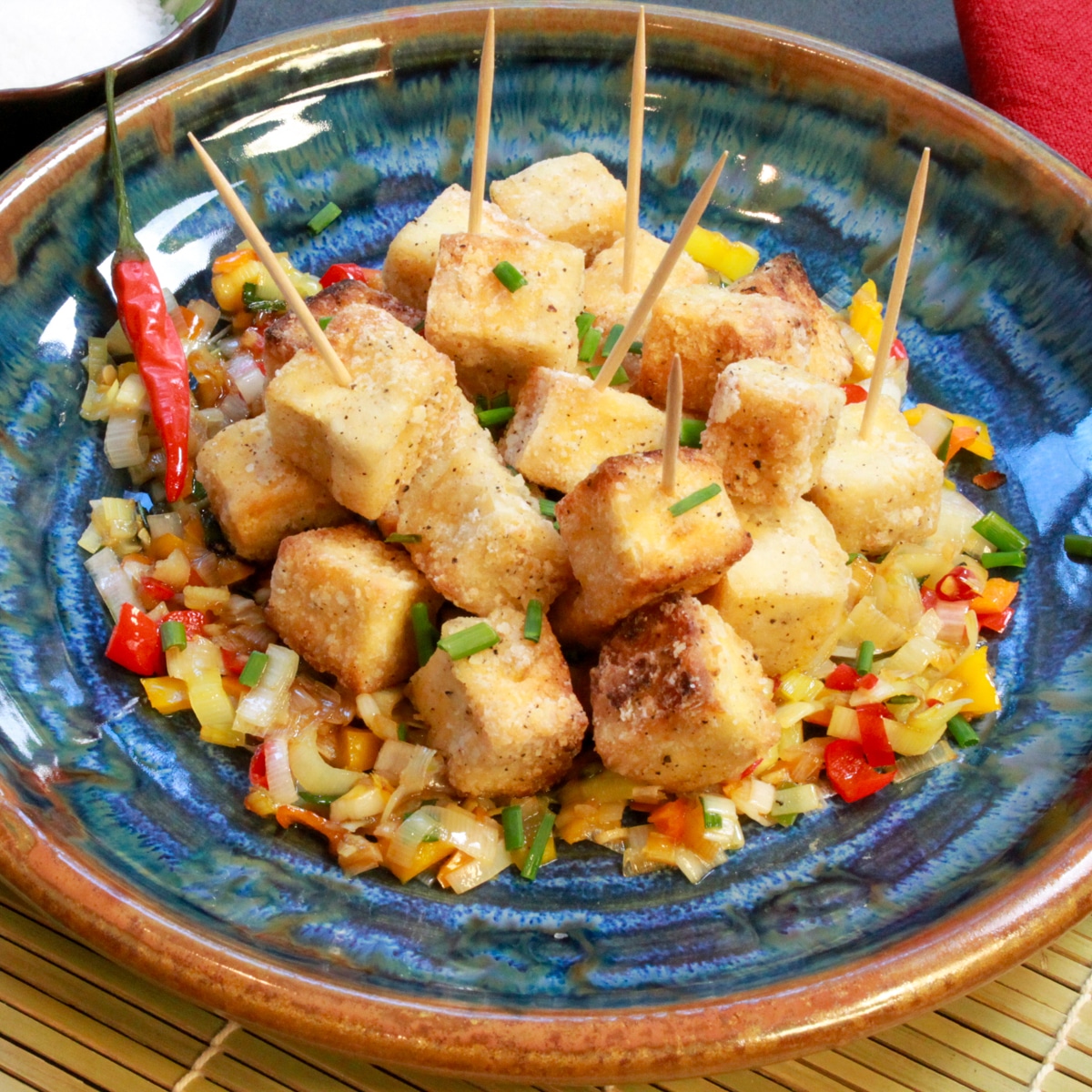 Crispy salt and pepper tofu cubes piled on top of sauteed vegetables on a round blue plate with a small bowl of salt on the side.