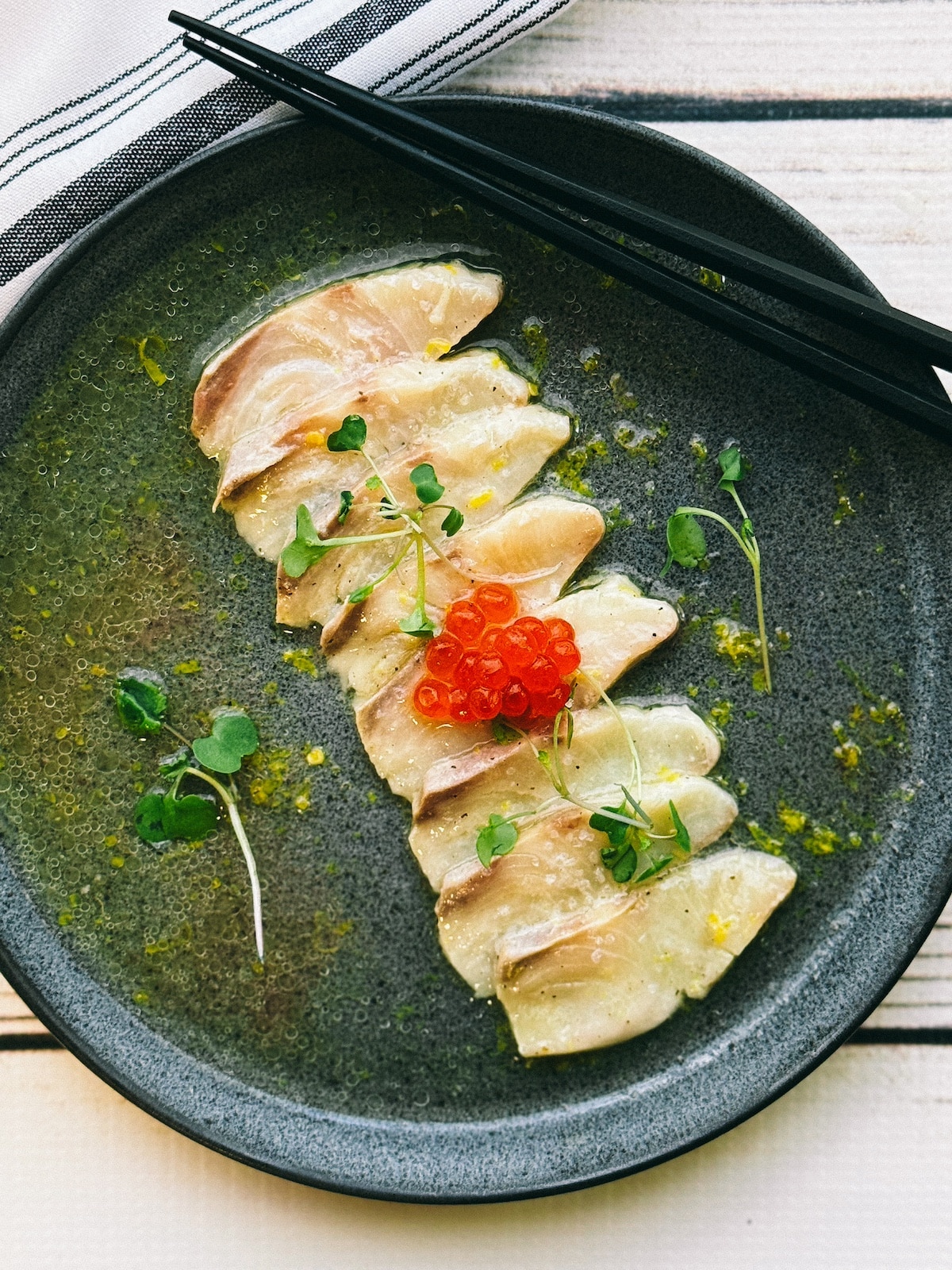 Fresh yellowtail slices topped with red fish eggs lined up on a round black plate on ponzu dressing on top of a white wooden board.