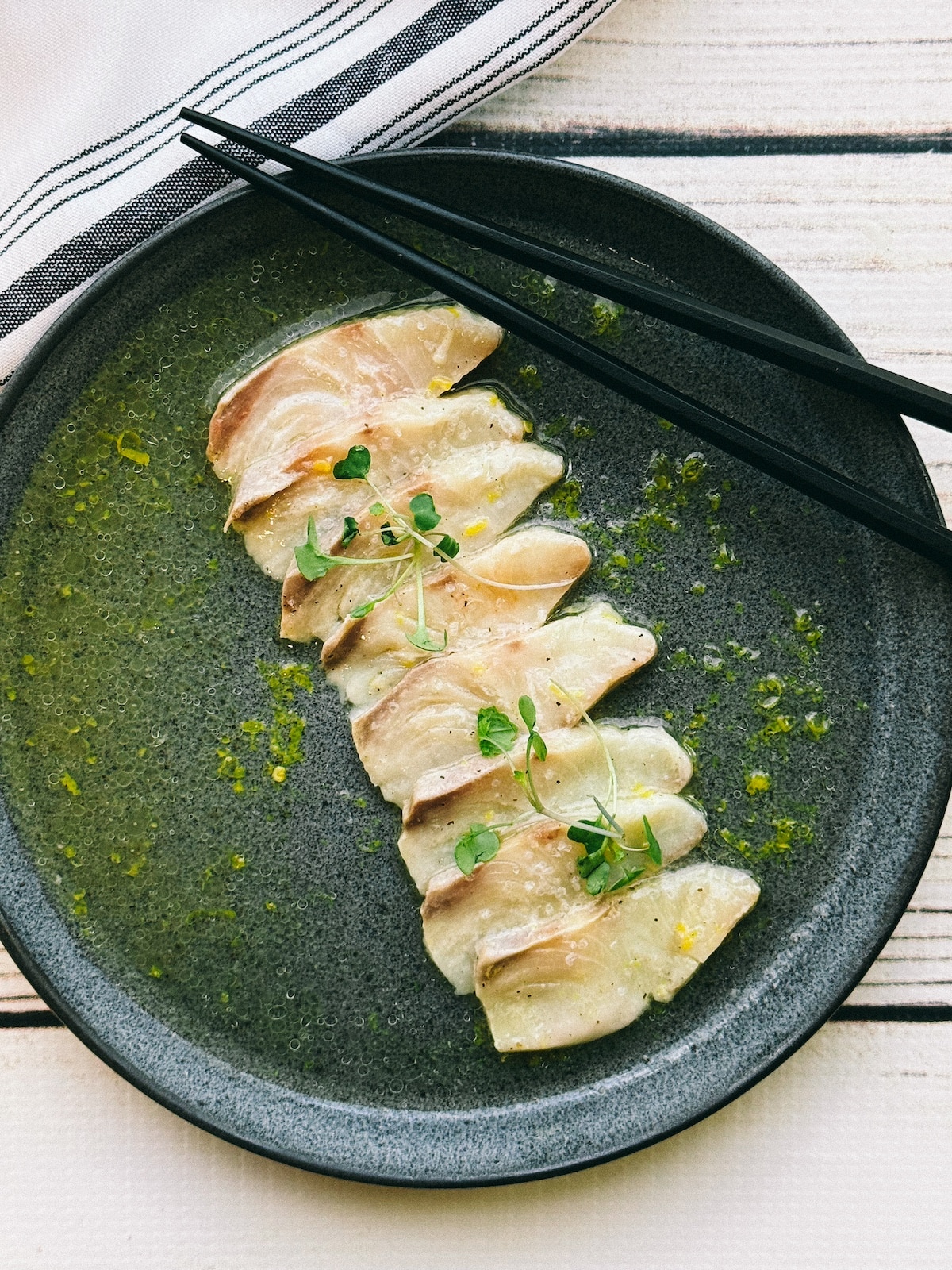 Fresh yellowtail slices lined up on a round black plate on ponzu dressing on top of a white wooden board.