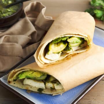 A grilled tofu and zucchini wrap sliced in half stacked on a blue plate with a napkin and sauce on the side, on top of a wooden board.