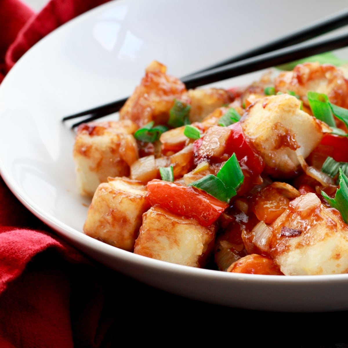 Crispy tofu cubes in a sweet and sour sauce on a round white plate with chopsticks, with a red napkin on the side.