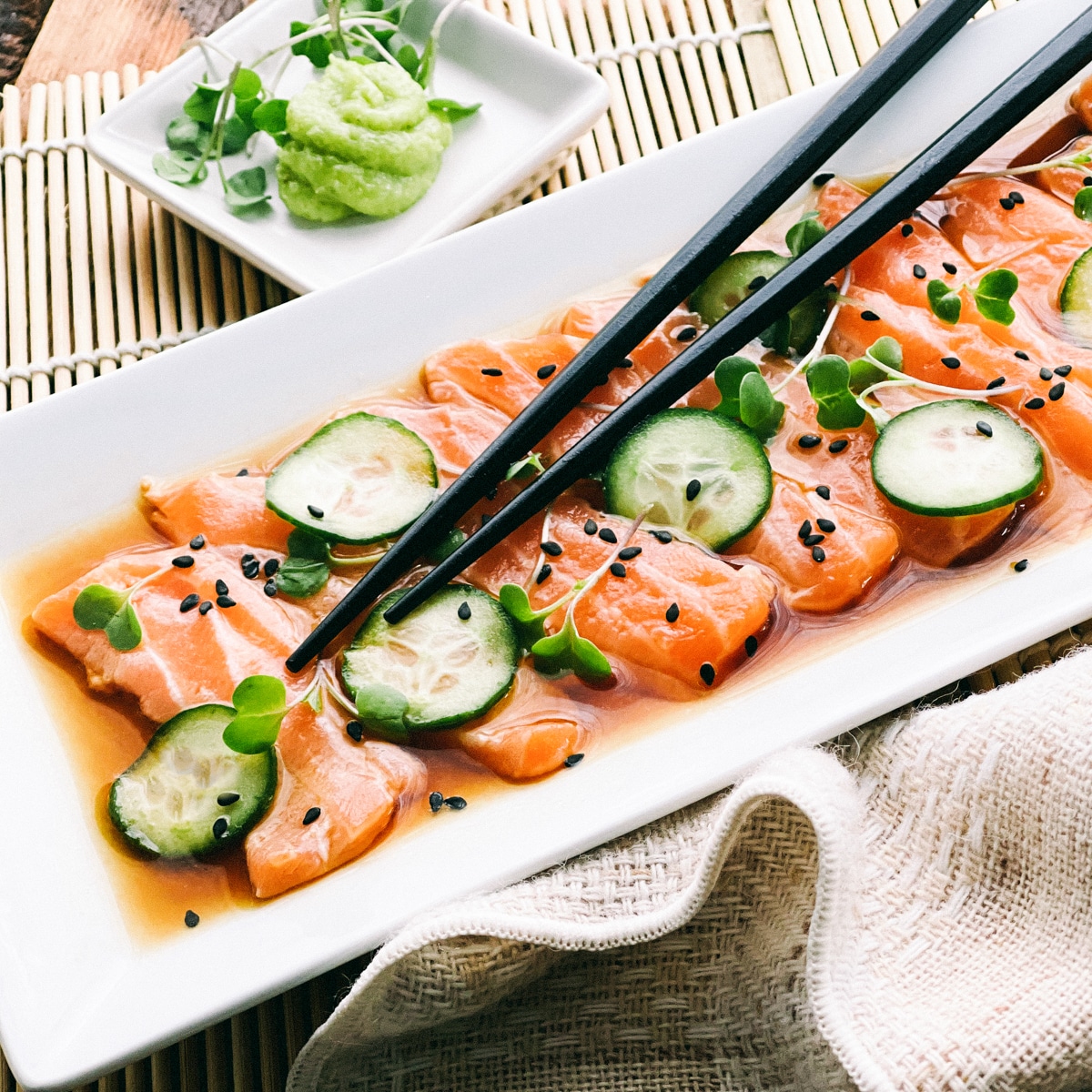 Slices of fresh sashimi salmon on a white platter topped with sliced cucumbers on top and sprinkles of black sesame seeds, with black chopsticks, a Japanese tea kettle, and a small white bowl of green wasabi on the side.