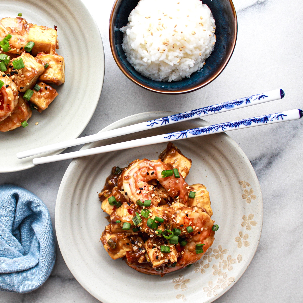 Two round white plates with General Tso's Shrimp and Tofu with a pair of blue and white chopsticks on top and a side of white rice, on a marble surface.