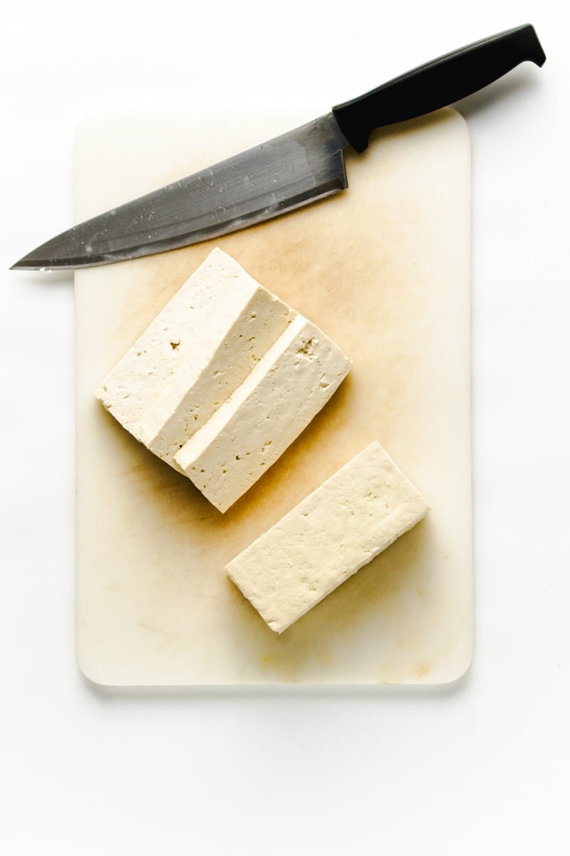 Slices of tofu on a white cutting board with a chef's knife on the side.