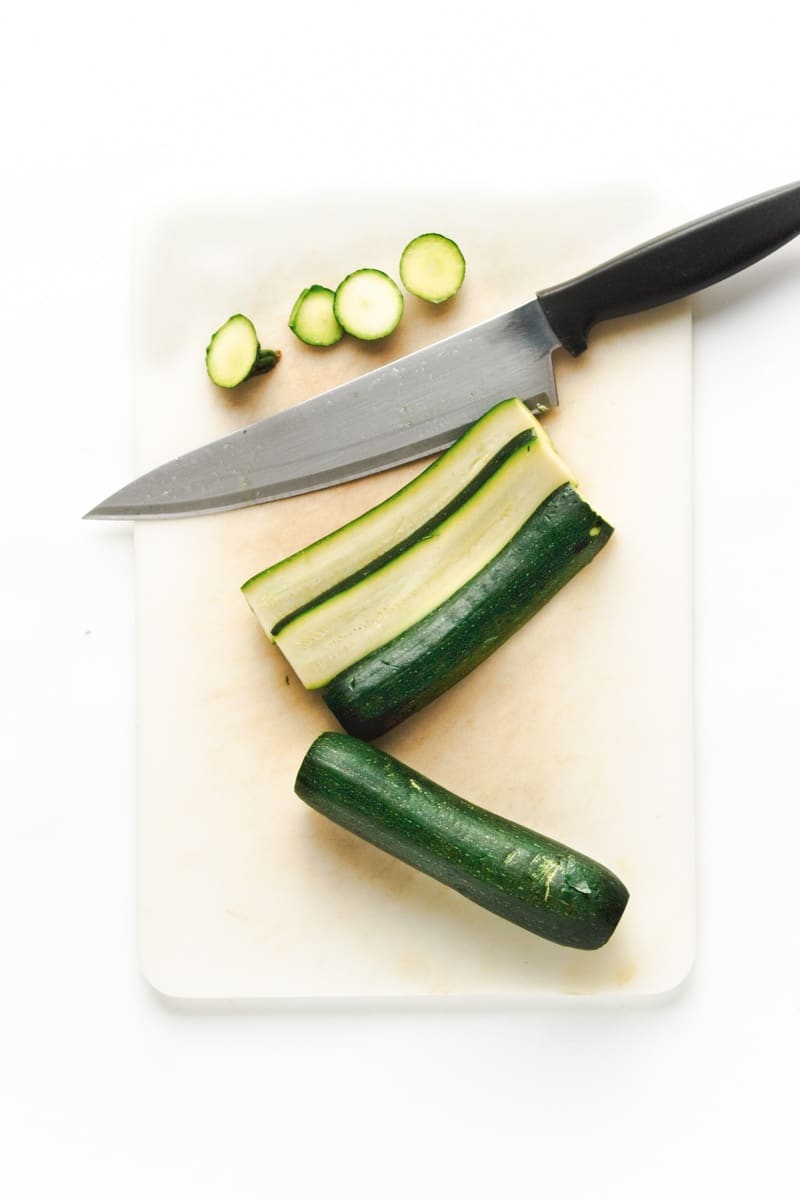 Sliced zucchini on a white cutting board with a chef's knife on the side.