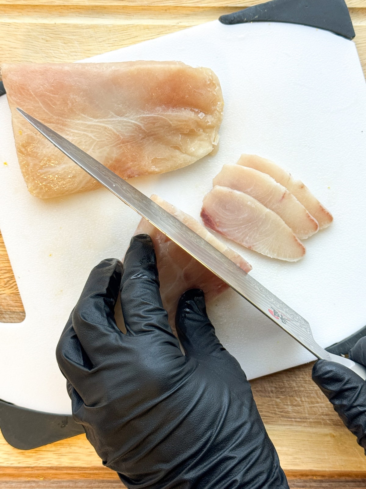 A black gloved hand slicing fresh hamachi yellowtail on top of white cutting board.