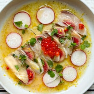 Hamachi yellowtail crudo on a white round plate with a yuzu lemon dressing surrounded by shaved radishes and topped with red fish eggs and microgreens.