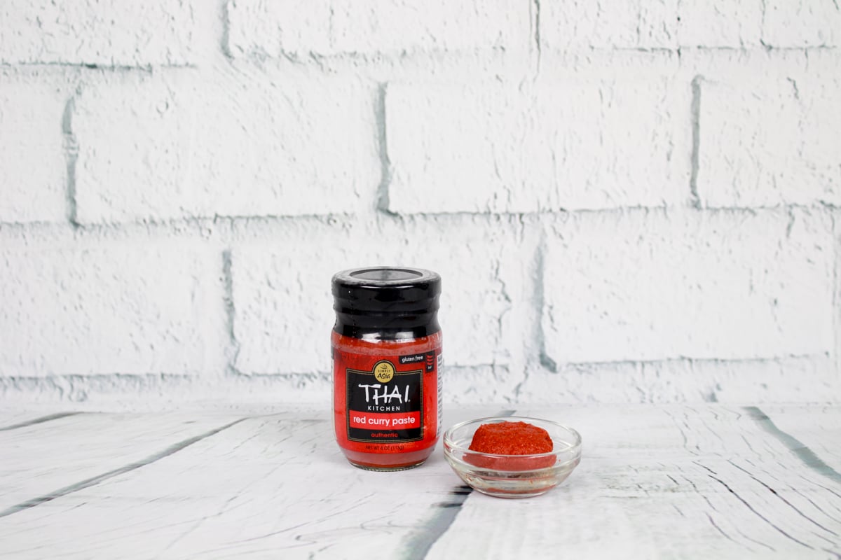 A bottle of Thai Kitchen red curry paste and a small glass bowl of red curry paste on top of white wooden planks with a white brick background.