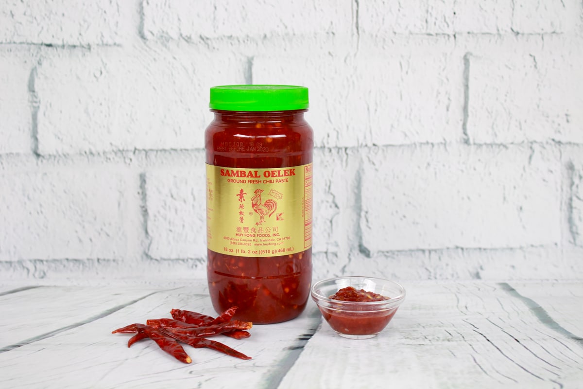 A bottle of sambal oelek and a small glass bowl filled with sambal oelek placed on top of a white plank board with white brick in the background.