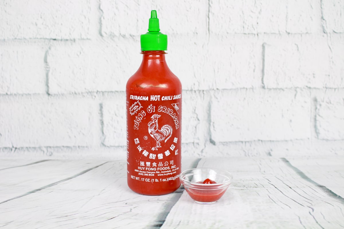 A bottle of Huy Fong Sriracha chili sauce and a small glass bowl filled with sriracha placed on top of a white plank board with white brick in the background.