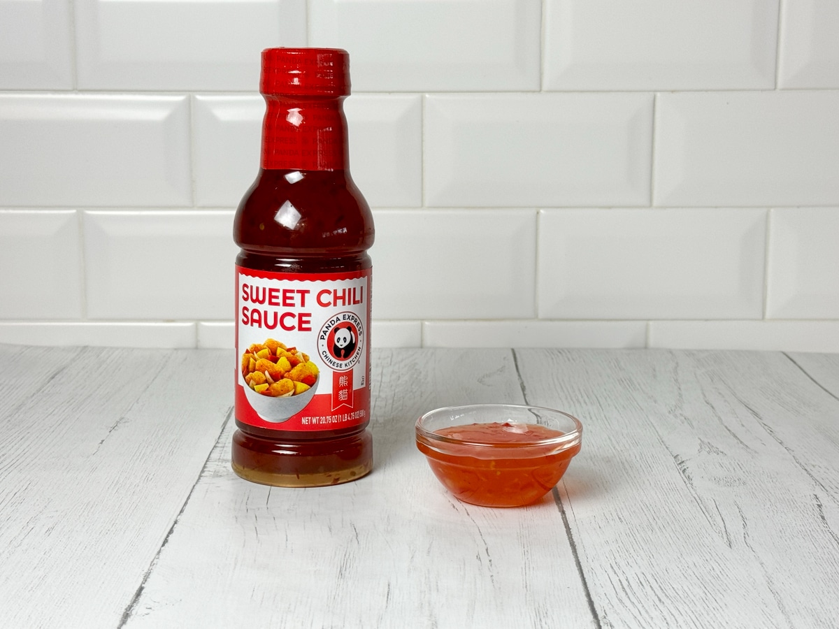 A bottle of Panda Express sweet chili sauce on top of white wooden planks with a white brick background.