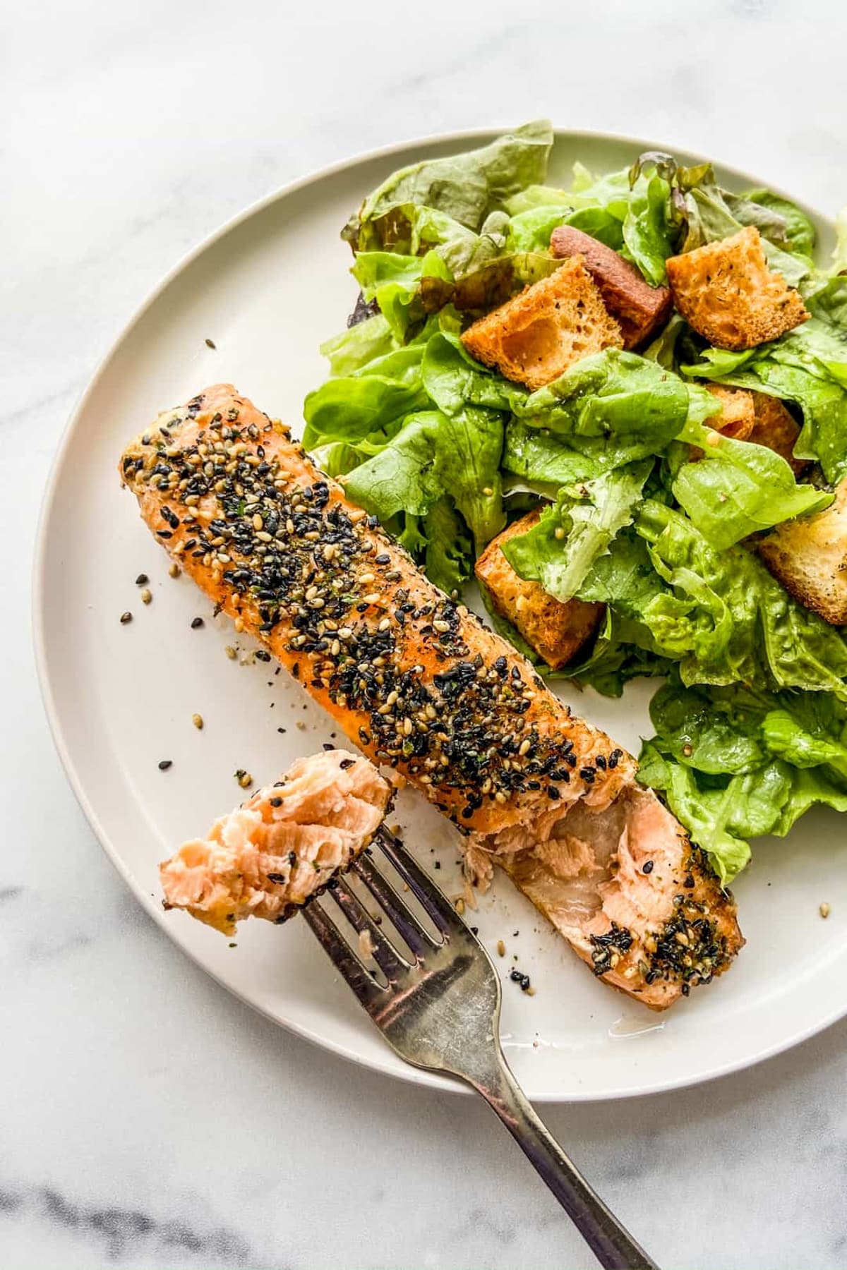 A healthy salmon recipe featuring a salmon filet topped with furikake seasoning on a round white plate with a side salad and a fork on top of a marble surface.