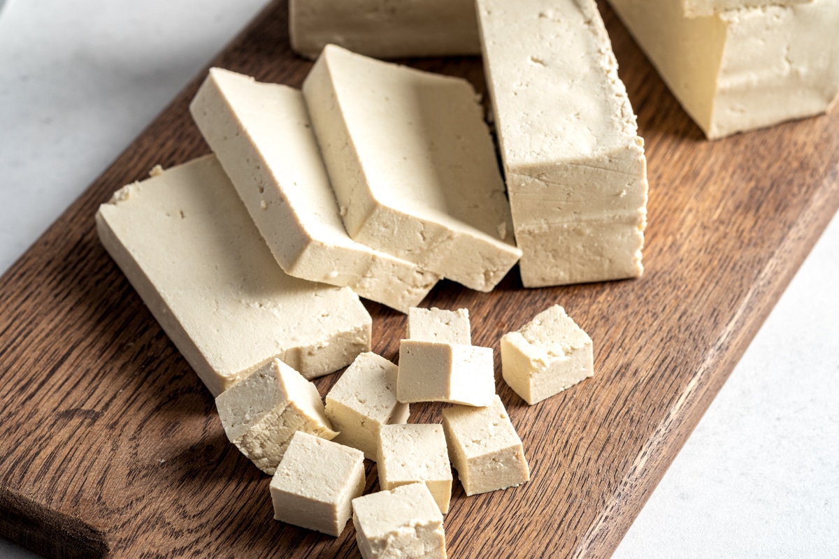 Firm tofu cut into slices and cubes on top of a wooden cutting board.