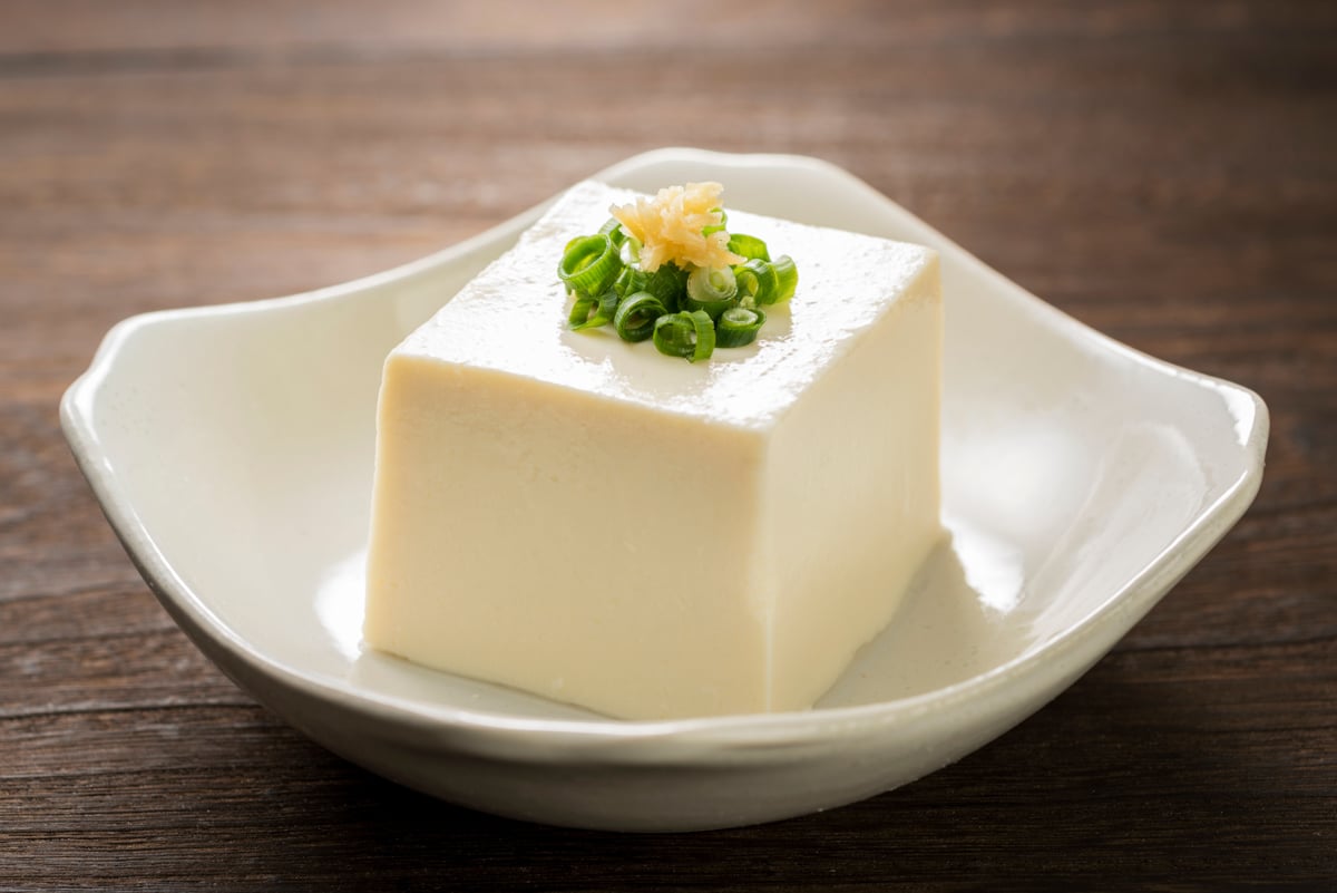 A block of silken tofu topped with sliced green onions in a white bowl on top of a wooden surface.
