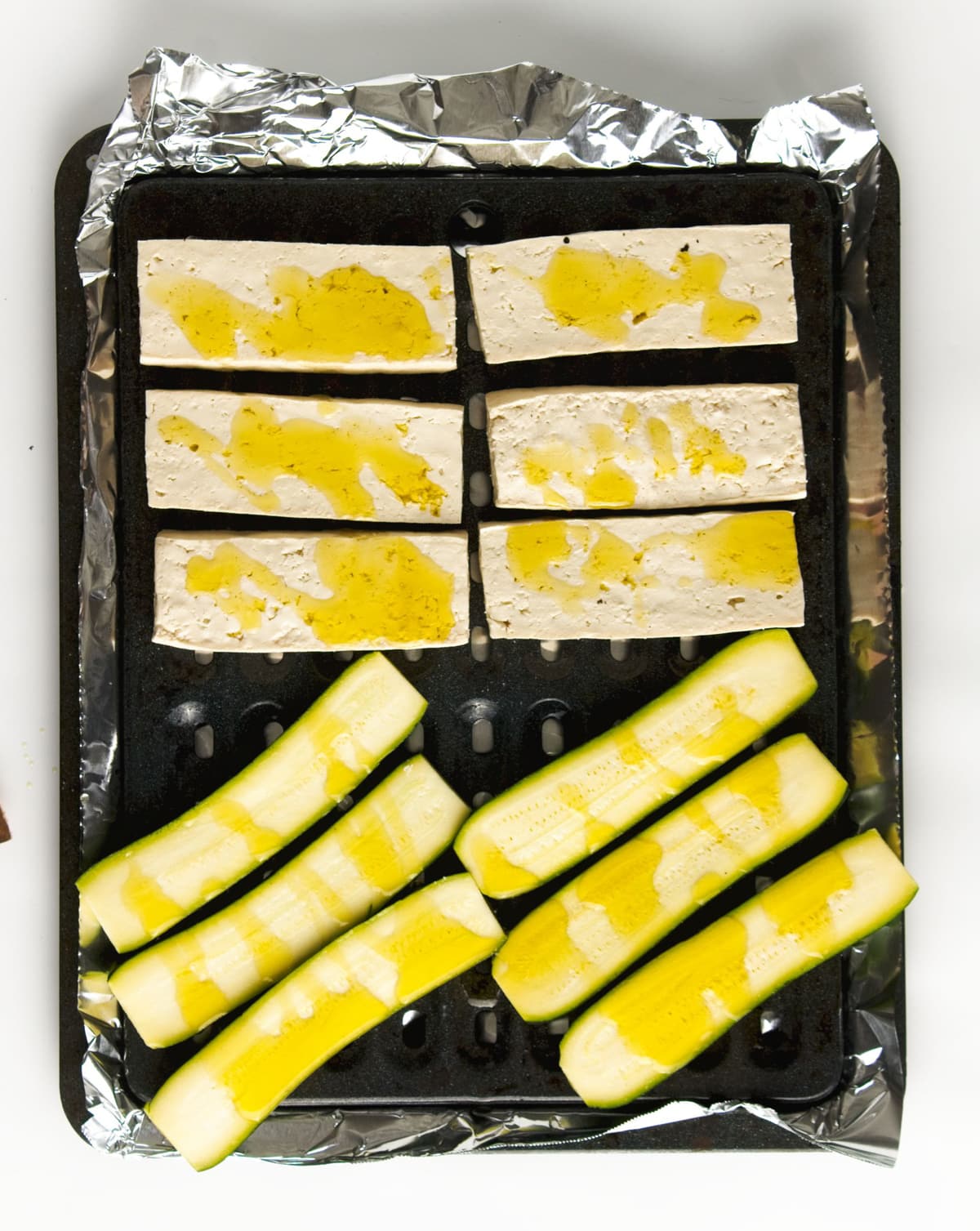 Tofu and zucchini strips brushed with oil on a black baking sheet lined with aluminum foil.