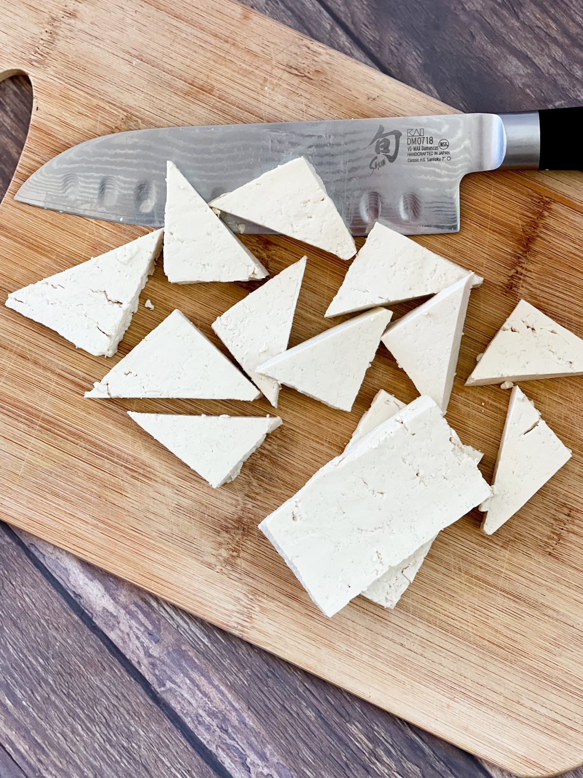 Tofu triangles being cut on a wooden cutting board with a chefs knife on the side.