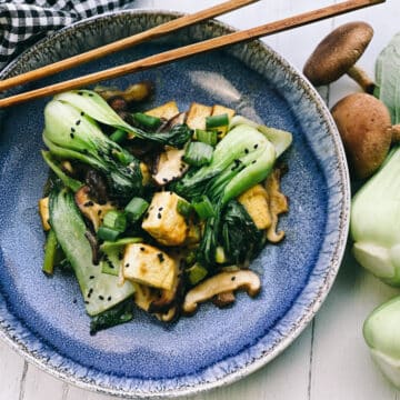 Stir-fry miso shiitake mushrooms, tofu, and baby bok choy on a blue plate with wooden chopsticks, and fresh shiitake mushrooms on the side.