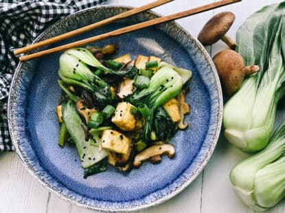 Stir-fry miso shiitake mushrooms, tofu, and baby bok choy on a blue plate with wooden chopsticks, and fresh shiitake mushrooms on the side.