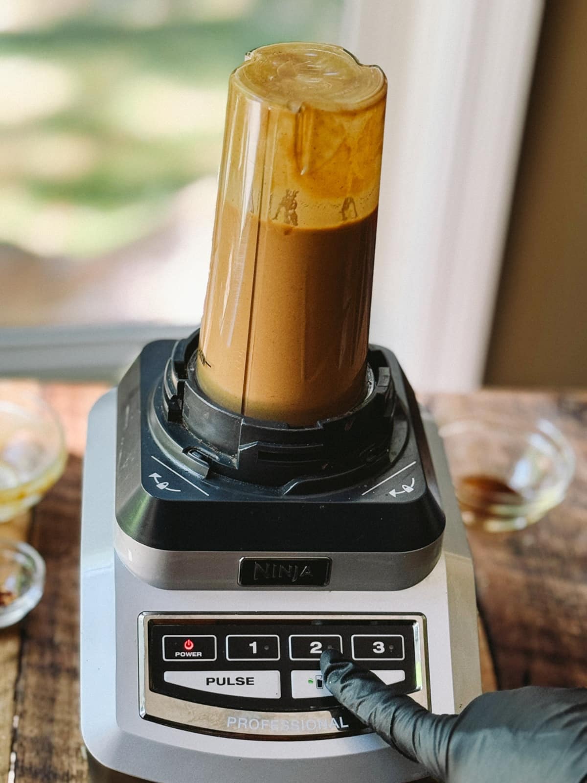 Ginger peanut dressing being blended in a tall clear glass on a blender, with a black gloved hand pushing the blend button on the blender.