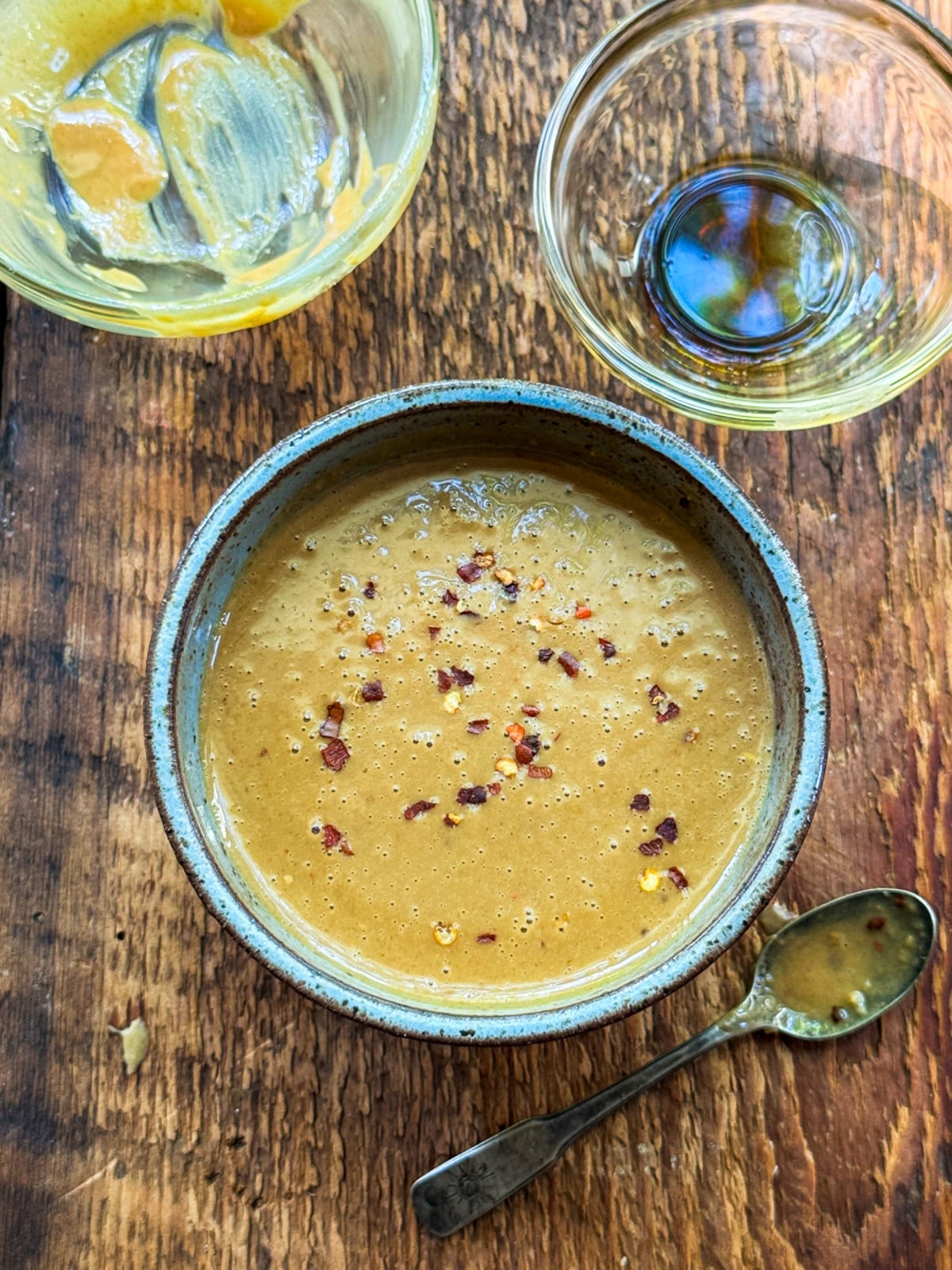 A round gray bowl filled with ginger peanut dressing topped with sprinkles of red pepper flakes, and a spoon and empty glass bowls on the side, on top of a wooden board.