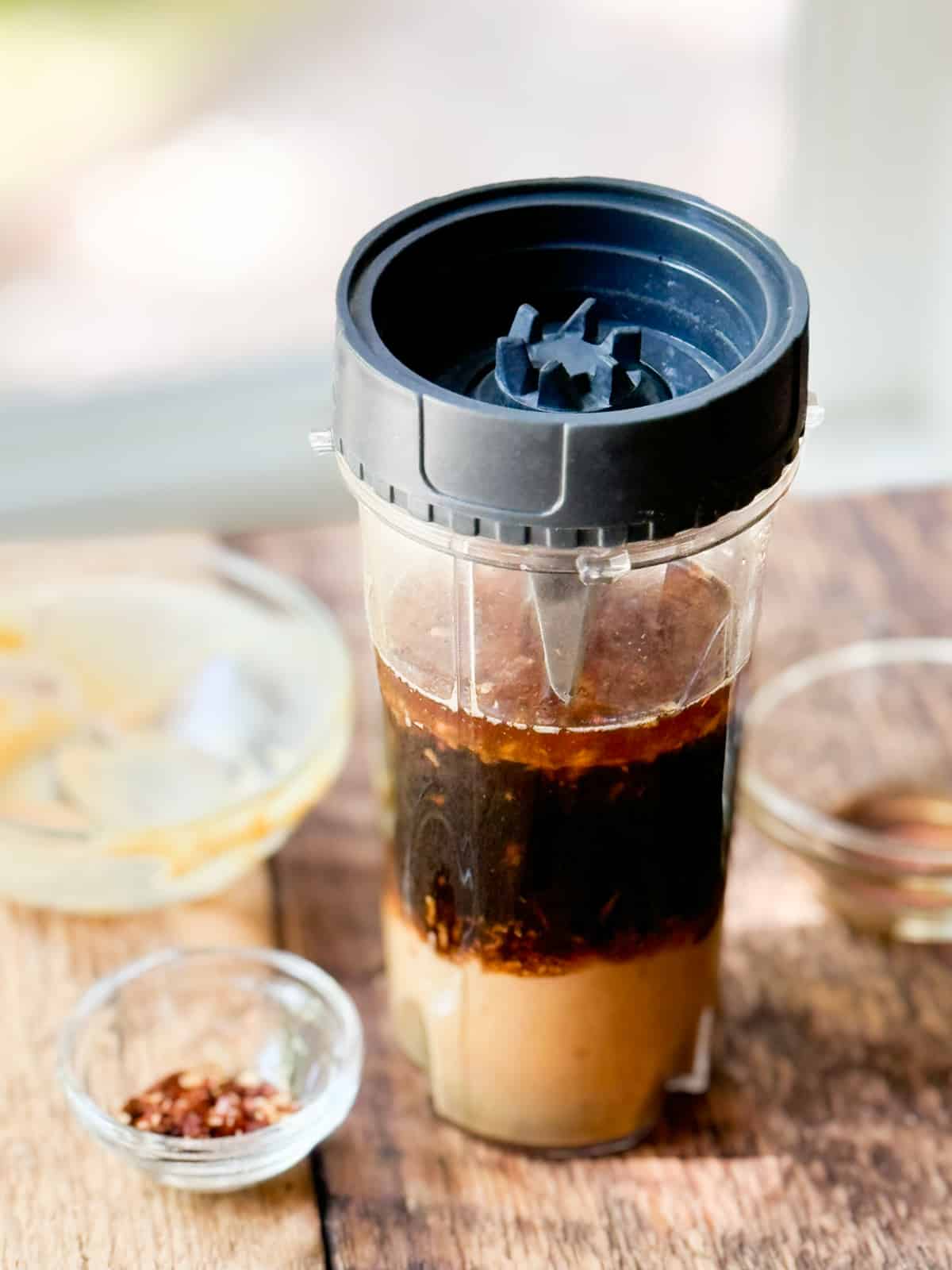 Unmixed ginger peanut dressing in layers in a tall clear glass with a black lid, and empty glass bowls on the side.