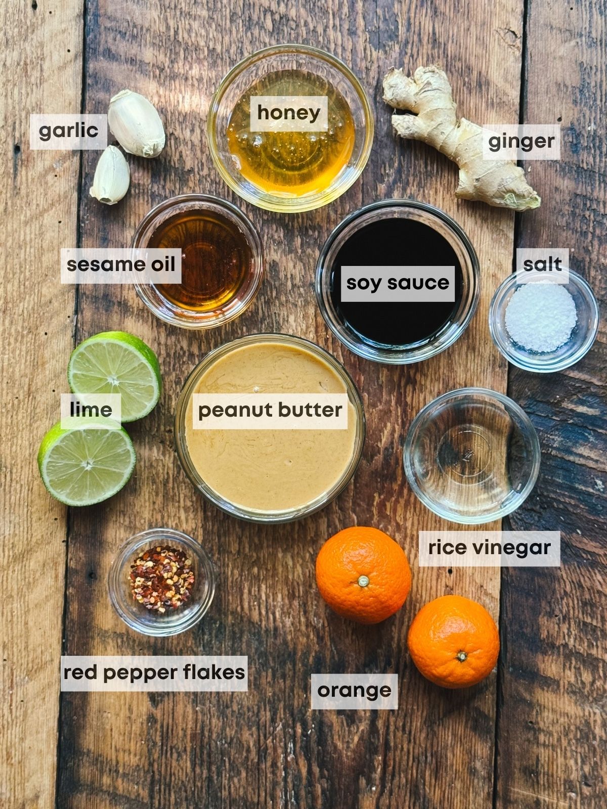 Labeled ingredients both in clear bowls and loose for making a ginger peanut dressing, placed on top of a wooden board.