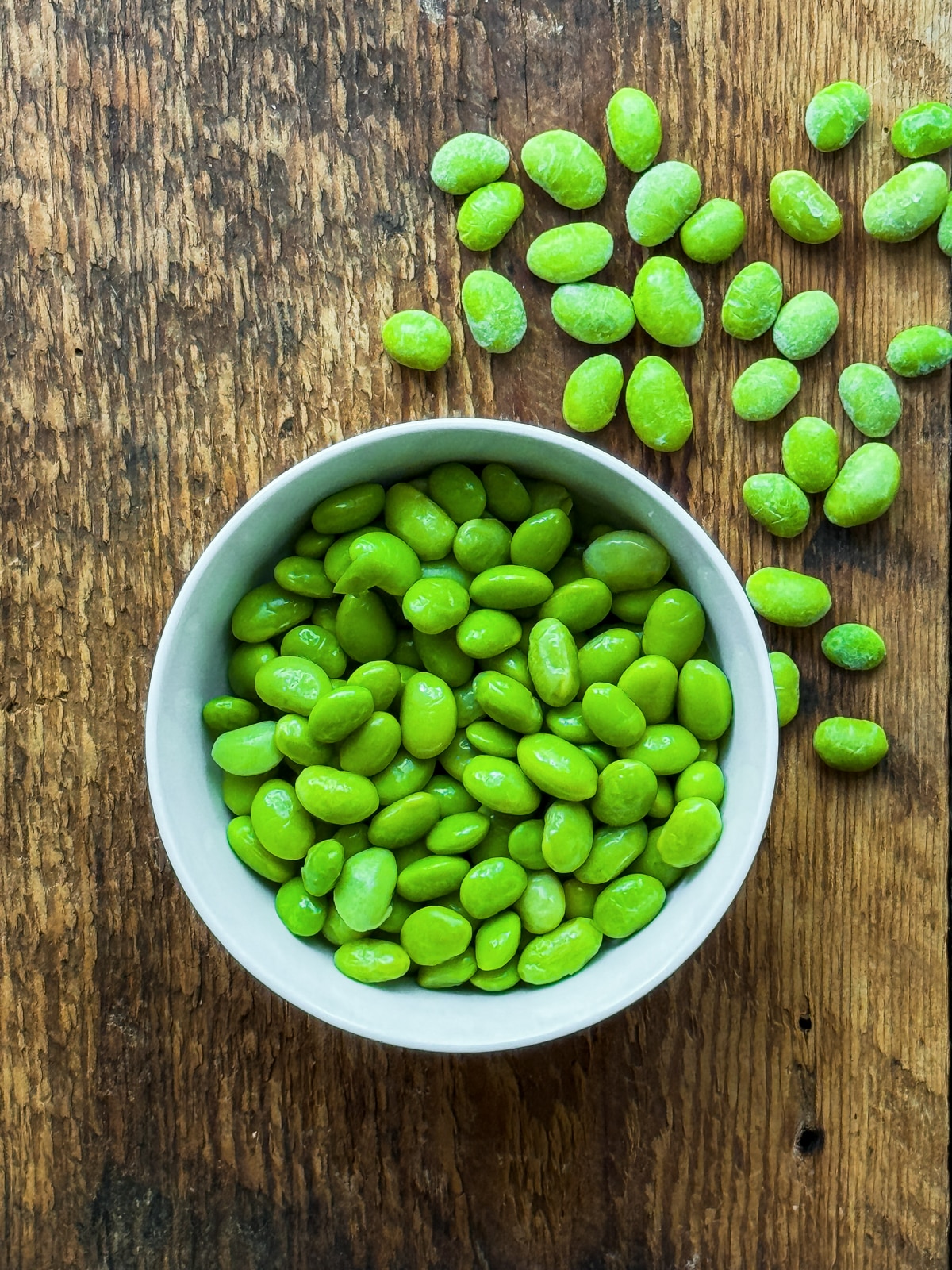 Shelled edamame in a white bowl on top of a wooden surface with scattered edamame on the side.