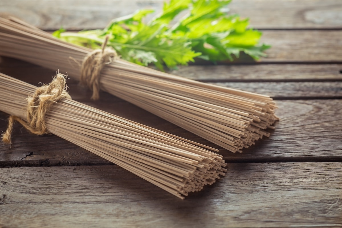 Two packets of soba noodles tied with string and placed on top of a wooden board with a sprig of green herbs in the background.