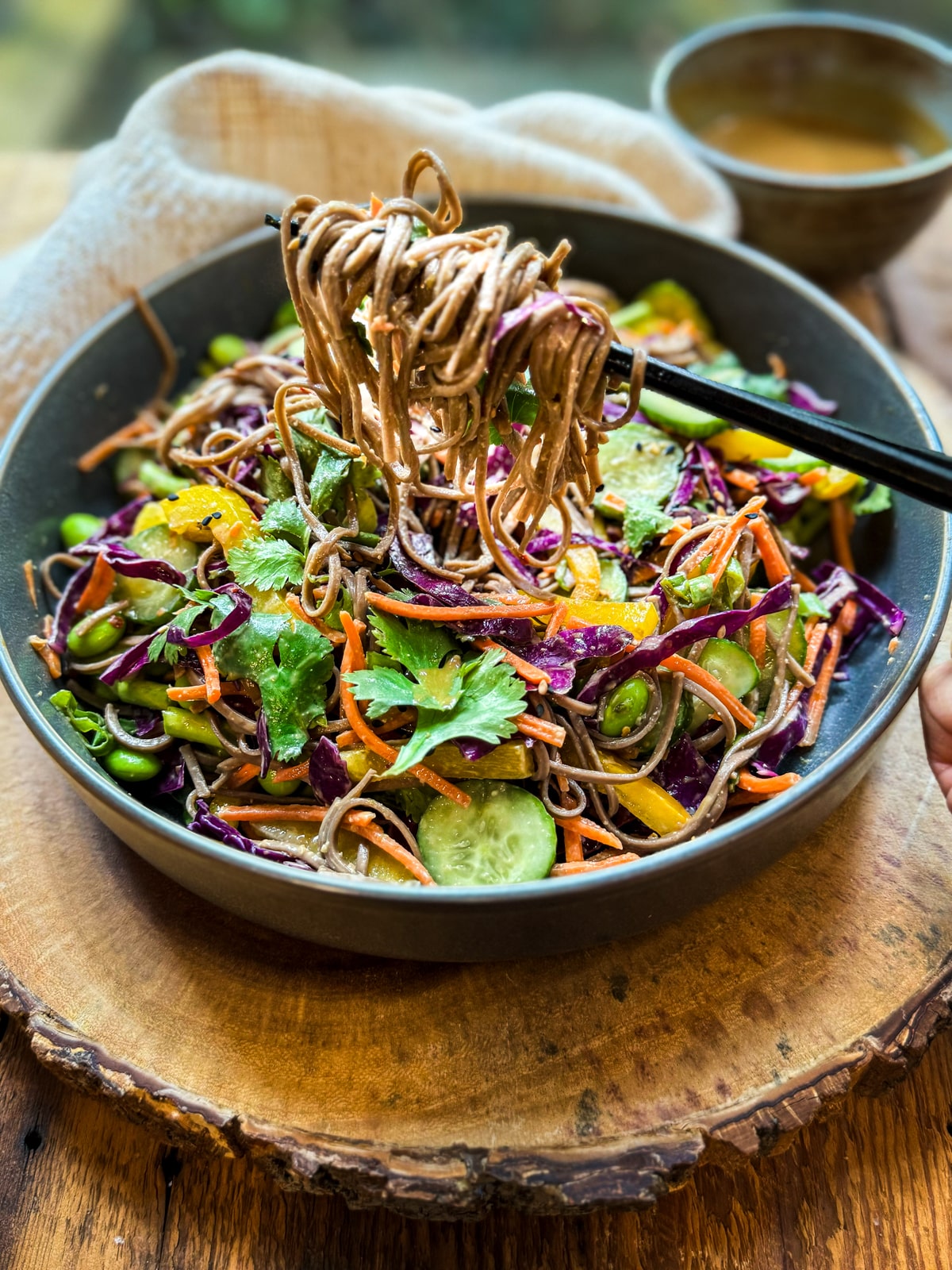 A vibrant Vegan Asian Noodle Salad in a dark gray bowl with soba noodles twirled on black chopsticks on a round wooden board with a linen napkin and small bowl of dressing on the side.