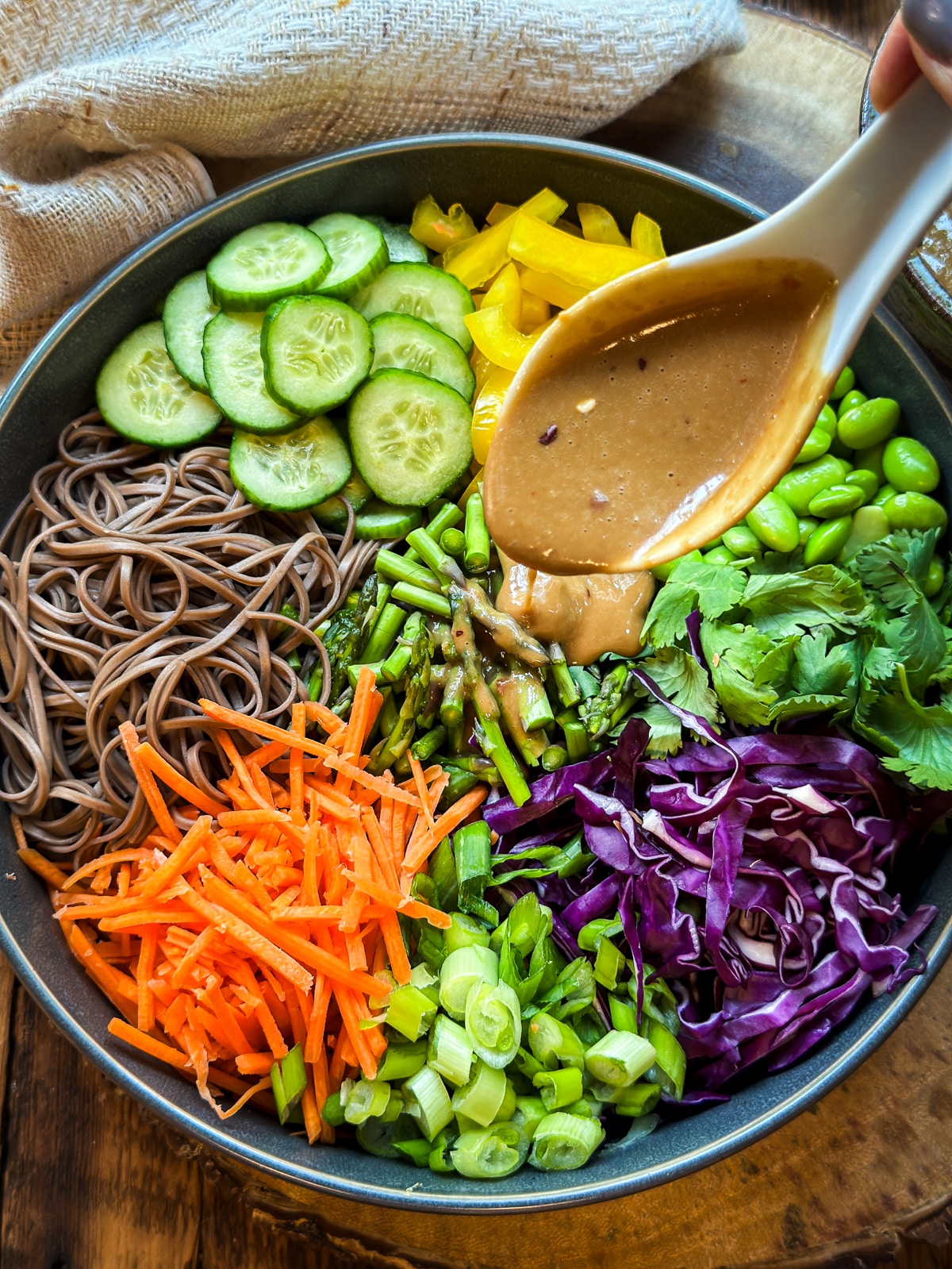 A white spoon pouring dressing over a vibrant Vegan Asian Noodle Salad in on dark gray bowl placed on top of a wooden board with a linen napkin on the side.