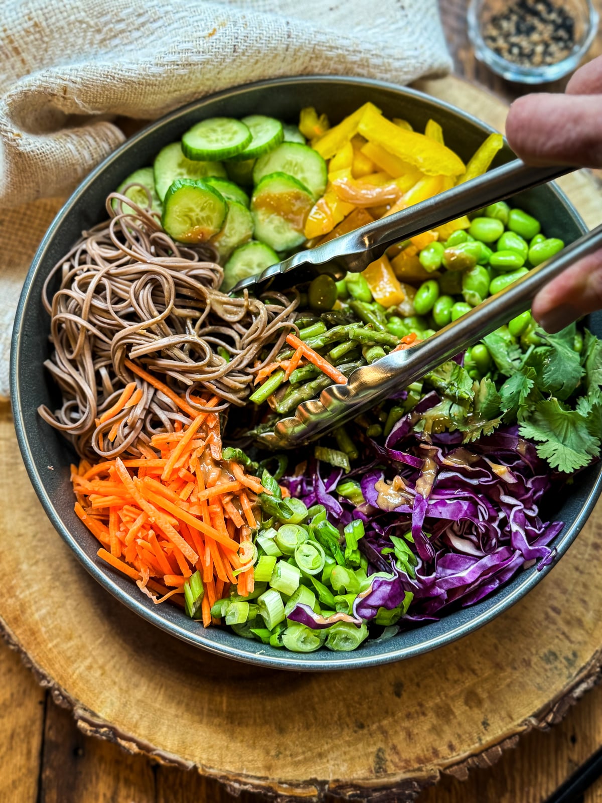 A pair of tongs mixing up a vibrant Vegan Asian Noodle Salad in a dark gray bowl placed on top of a wooden board with a linen napkin and a small bowl of sesame seeds on the side.