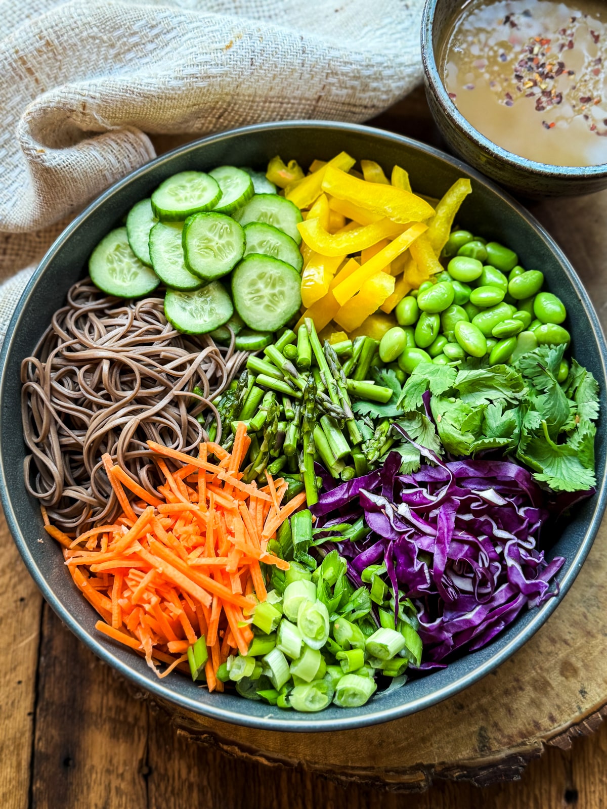 Vibrant, crisp vegetables and soba noodles in a dark gray bowl ready for mixing up a Vegan Asian Noodle Salad placed on top of a wooden board with a linen napkin and a small bowl of dressing on the side.