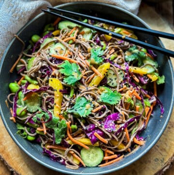 A vibrant Vegan Asian Noodle Salad in a dark gray bowl topped with black chopsticks on a round wooden board with a linen napkin and small bowl of dressing on the side.