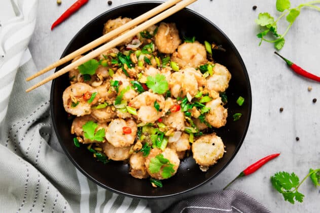 Chinese salt and pepper shrimp in a black bowl with sautéed vegetables and herbs, and a pair of chopsticks on top on a gray surface with a gray