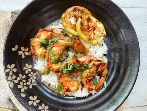 Grilled shrimp with honey garlic miso on top of white rice in a black bowl with grilled lemon on the side.