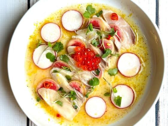 Hamachi yellowtail crudo on a white round plate with a yuzu lemon dressing surrounded by shaved radishes and topped with red fish eggs and microgreens.