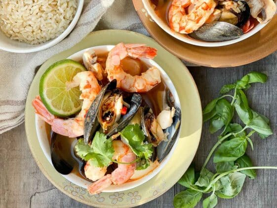 A white bowl of Thai seafood soup with shrimp, mussels and lime on top of a yellow plate with fresh herbs and a small bowl of rice on the side placed on top of a wooden board.