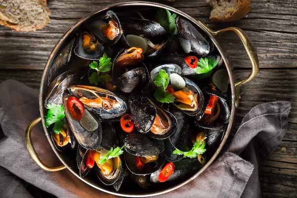 An overhead shot of a copper pot filled with steamed mussels garnished with red chili peppers and cilantro on a wooden board with sliced bread and a gray napkin on the side.