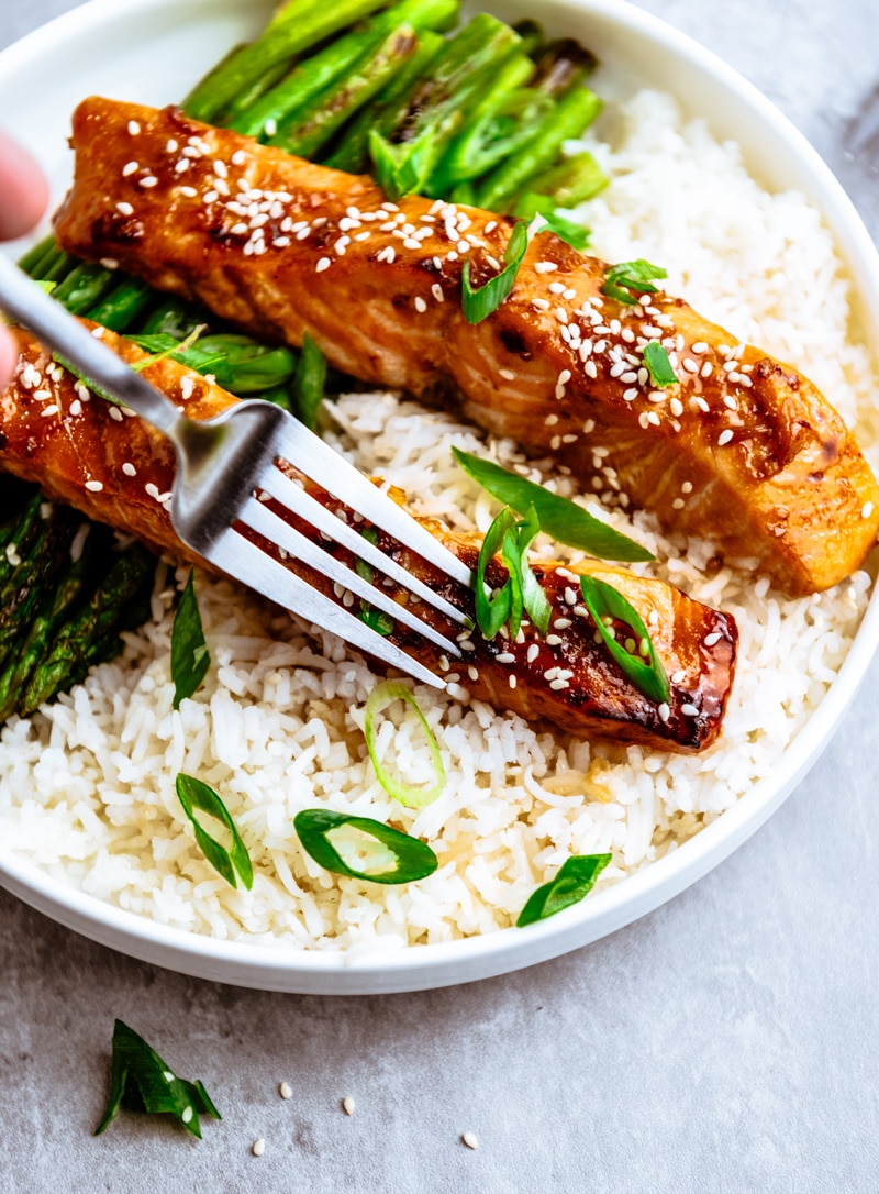 Air Fryer Teriyaki Salmon on top of white rice with fork on a white round plate with roasted asparagus on the side.