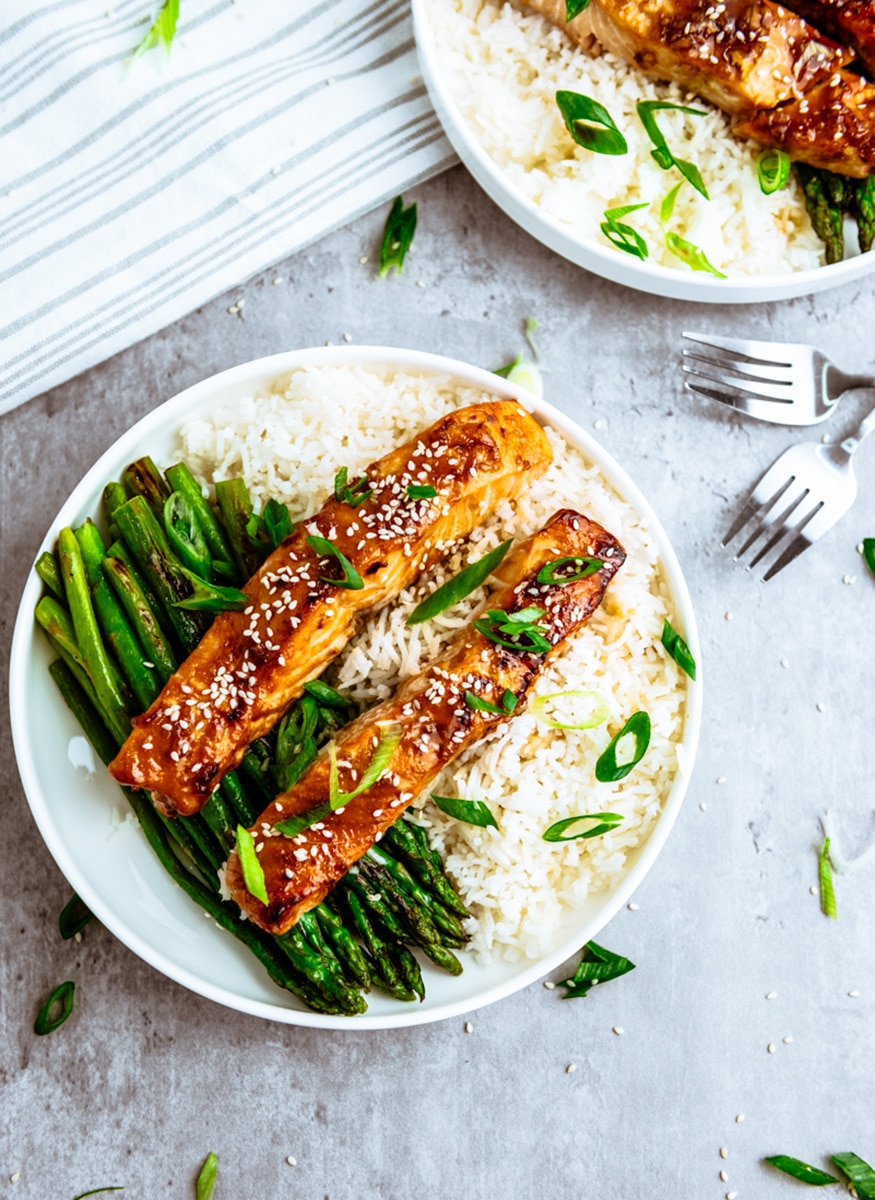 Two round plates of Air Fryer Teriyaki Salmon on top of white rice with roasted asparagus on the side, placed on a gray surface with silver forks.