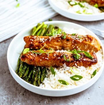 Air Fryer Teriyaki Salmon on top of white rice with fork on a white round plate with roasted asparagus on the side.