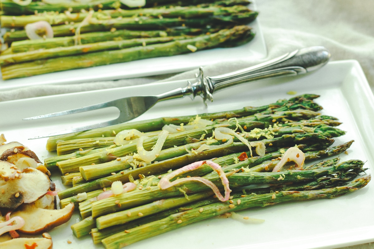 Roasted asparagus and shiitake mushrooms on a white rectangular plate with a silver aspargus fork on the side.