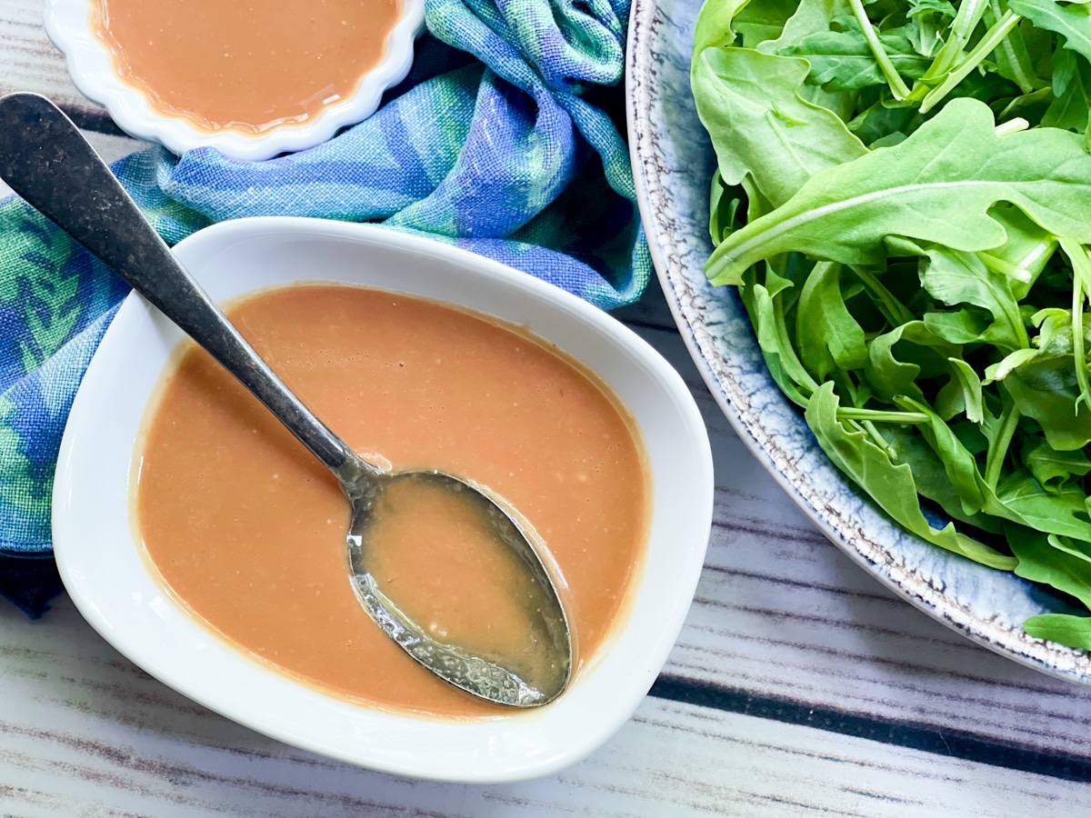 Miso dressing in a small white bowl with a spoon inserted and a bowl of greens on the side with a blue napkin.